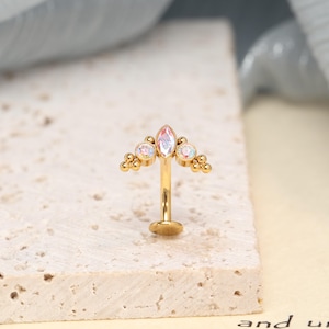 Puede incluir: Una joya corporal de barra curva en tono dorado con una parte superior decorativa que presenta una piedra central iridiscente de corte marquesa, flanqueada por dos piedras redondas iridiscentes y pequeñas esferas doradas.