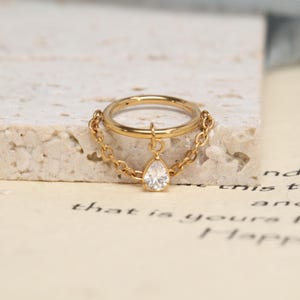 May include: A gold-toned hoop ring featuring a delicate chain draped across the front. A single pear-shaped clear gemstone charm hangs from the center of the chain, suspended from the hoop by a small jump ring. The metal has a polished finish.