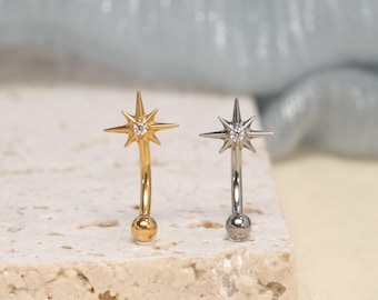 Könnte beinhalten: Zwei gebogene Piercing-Stäbe mit einem Sternmotiv an der Oberseite. Ein Stab ist goldfarben mit einem klaren Mittelstein, der andere ist silberfarben mit einem klaren Mittelstein. Jedes Schmuckstück hat eine kleine runde Kugel am unteren Ende.