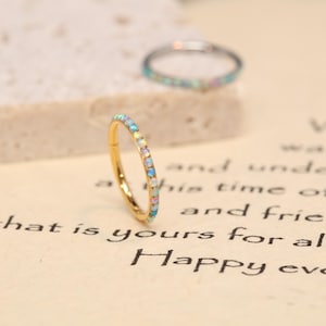 May include: A gold-colored ring with a row of small, iridescent opal stones. The ring is in focus, with a second ring slightly blurred in the background. The background is a light-colored surface with printed text.