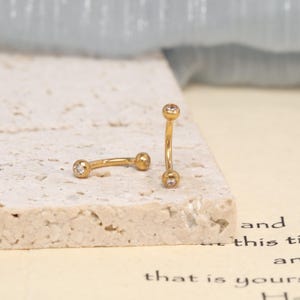 K&ouml;nnte beinhalten: Zwei goldfarbene, gebogene Barbell-Piercings, die jeweils an beiden Enden mit einem kleinen, klaren, rund geschliffenen Edelstein in der Kugel besetzt sind. Ein Schmuckst&uuml;ck liegt horizontal, w&auml;hrend das andere vertikal aufrecht steht.