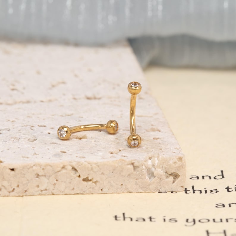 K&ouml;nnte beinhalten: Zwei goldfarbene, gebogene Barbell-Piercings, die jeweils an beiden Enden mit einem kleinen, klaren, rund geschliffenen Edelstein in der Kugel besetzt sind. Ein Schmuckst&uuml;ck liegt horizontal, w&auml;hrend das andere vertikal aufrecht steht.