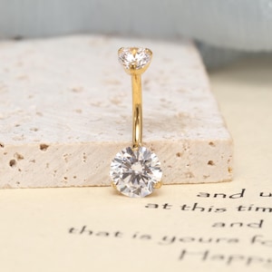May include: A gold-toned curved barbell belly button ring featuring a small round clear gemstone at the top and a larger round clear gemstone at the bottom. The metal post is polished and smooth, connecting the two faceted stones securely.
