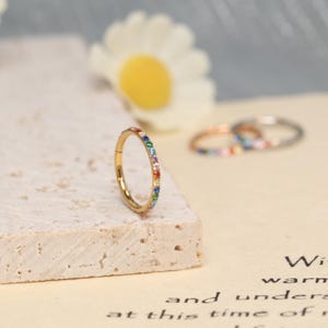 May include: A gold-colored hoop earring with a row of colorful gemstones. The earring is set against a neutral background, with a blurred daisy and other earrings in the background. The gemstones are in a rainbow pattern.