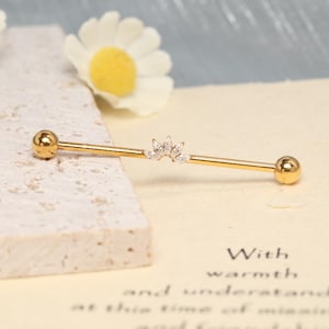 May include: A gold-toned industrial barbell piercing with a smooth, straight metal bar and two spherical end caps. The centre of the bar features a decorative arch-shaped attachment adorned with five small, clear, marquise-cut gemstones.