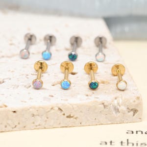 Peut inclure: Une collection de huit bijoux de piercing labret disposés sur deux rangées. La rangée supérieure présente quatre tiges en métal argenté, tandis que la rangée inférieure comporte quatre tiges dorées. Chaque bijou est orné d'une pierre opale.
