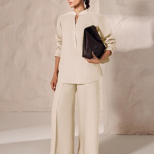 May include: A cream-colored linen outfit consisting of a long-sleeved tunic and wide-leg pants. The outfit is paired with a dark brown, textured leather handbag and brown sandals. The tunic has a mandarin collar and a front placket.