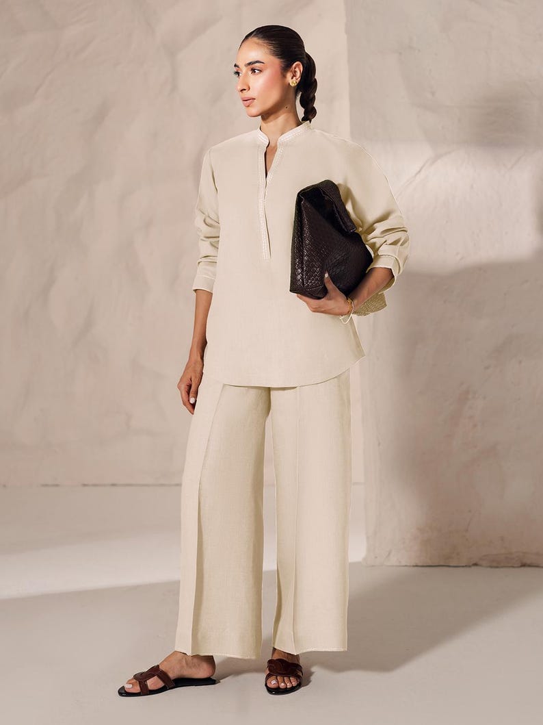 May include: A cream-colored linen outfit consisting of a long-sleeved tunic and wide-leg pants. The outfit is paired with a dark brown, textured leather handbag and brown sandals. The tunic has a mandarin collar and a front placket.
