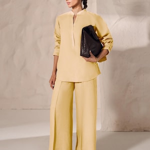 May include: A woman's ensemble featuring a pale yellow tunic with a mandarin collar and wide-leg pants. She holds a dark brown, woven leather handbag. The outfit is paired with brown sandals, creating a monochromatic look.