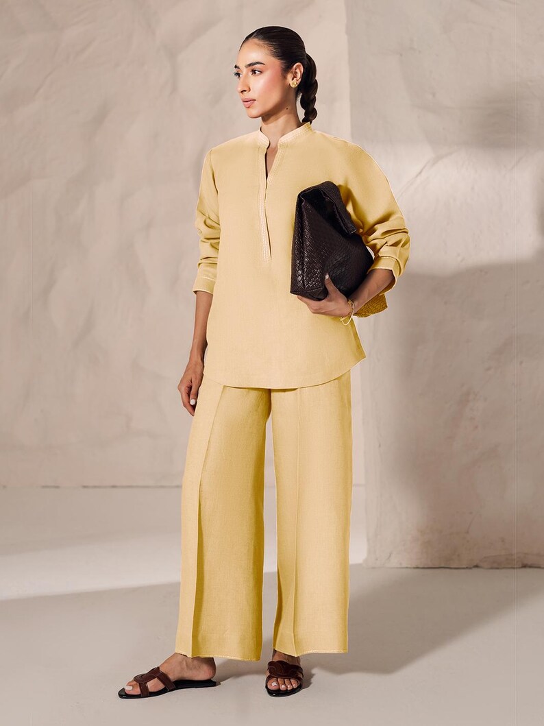 May include: A woman's ensemble featuring a pale yellow tunic with a mandarin collar and wide-leg pants. She holds a dark brown, woven leather handbag. The outfit is paired with brown sandals, creating a monochromatic look.