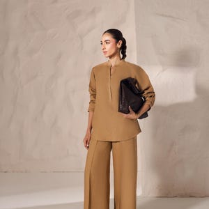 May include: A woman wearing a tan-colored outfit, including a long-sleeved tunic and wide-leg pants. She is holding a dark brown, woven leather tote bag. The outfit is paired with brown sandals. The background is a textured, light-colored wall.
