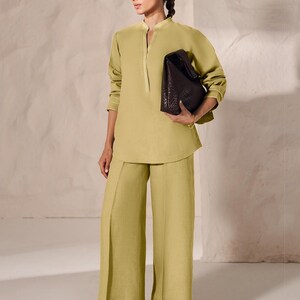 May include: A woman wearing a light green linen outfit, consisting of a long-sleeved tunic and wide-leg pants. The tunic has a mandarin collar and gold trim. She carries a dark brown, textured leather bag and wears brown sandals.