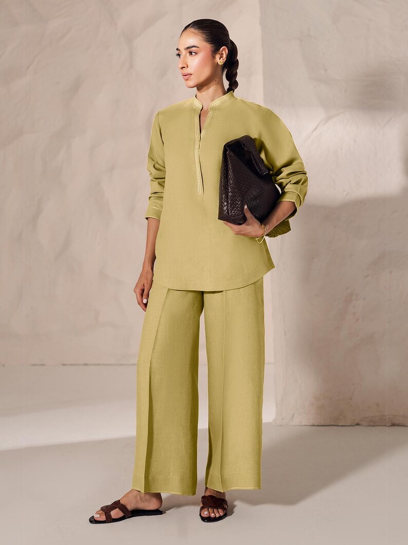May include: A woman wearing a light green linen outfit, consisting of a long-sleeved tunic and wide-leg pants. The tunic has a mandarin collar and gold trim. She carries a dark brown, textured leather bag and wears brown sandals.