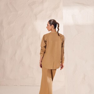 May include: A tan-colored two-piece outfit, consisting of a long-sleeved button-up shirt and wide-leg pants. The shirt has a mandarin collar and the pants have a high waist. The outfit is made of a smooth, possibly linen-like fabric.