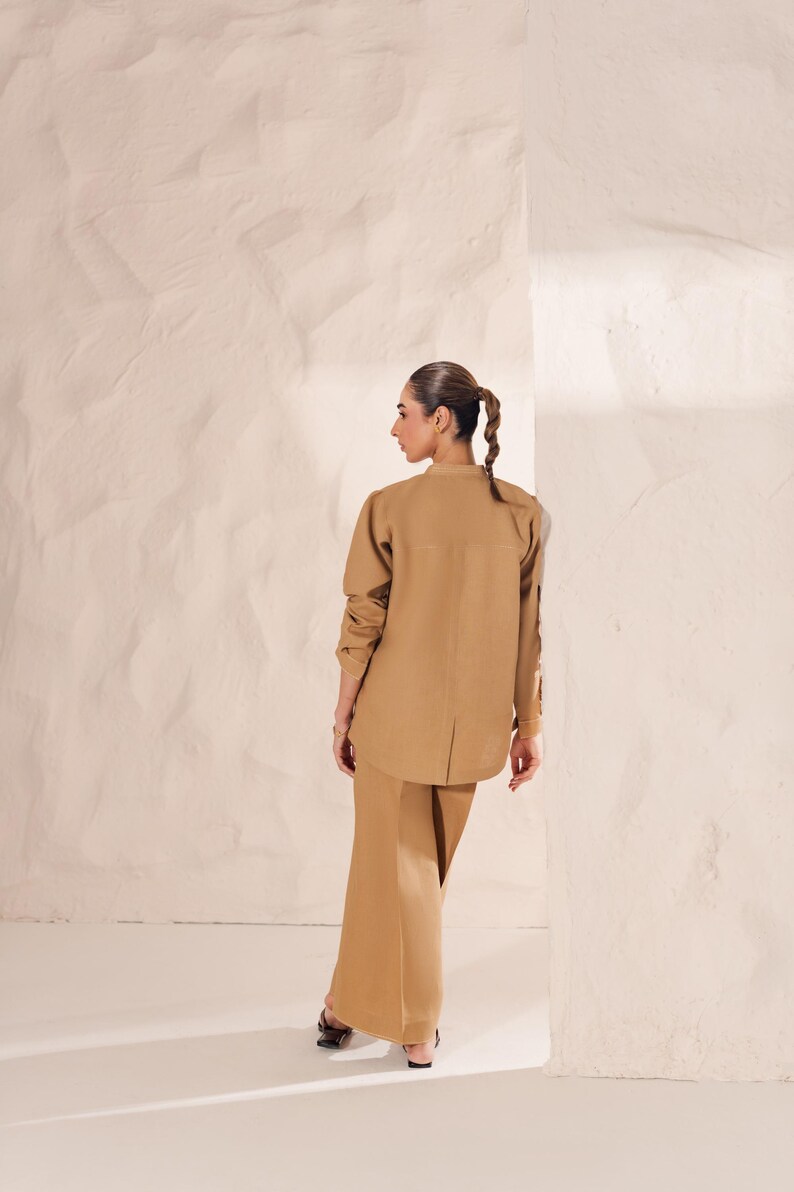 May include: A tan-colored two-piece outfit, consisting of a long-sleeved button-up shirt and wide-leg pants. The shirt has a mandarin collar and the pants have a high waist. The outfit is made of a smooth, possibly linen-like fabric.