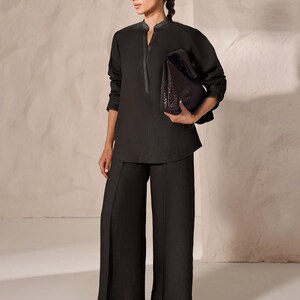 May include: A dark charcoal-colored ensemble featuring a long-sleeved tunic with a mandarin collar and wide-leg pants. The tunic has decorative stitching along the collar and cuffs. A dark brown leather tote bag is held in the hand. The outfit is paired with brown sandals.