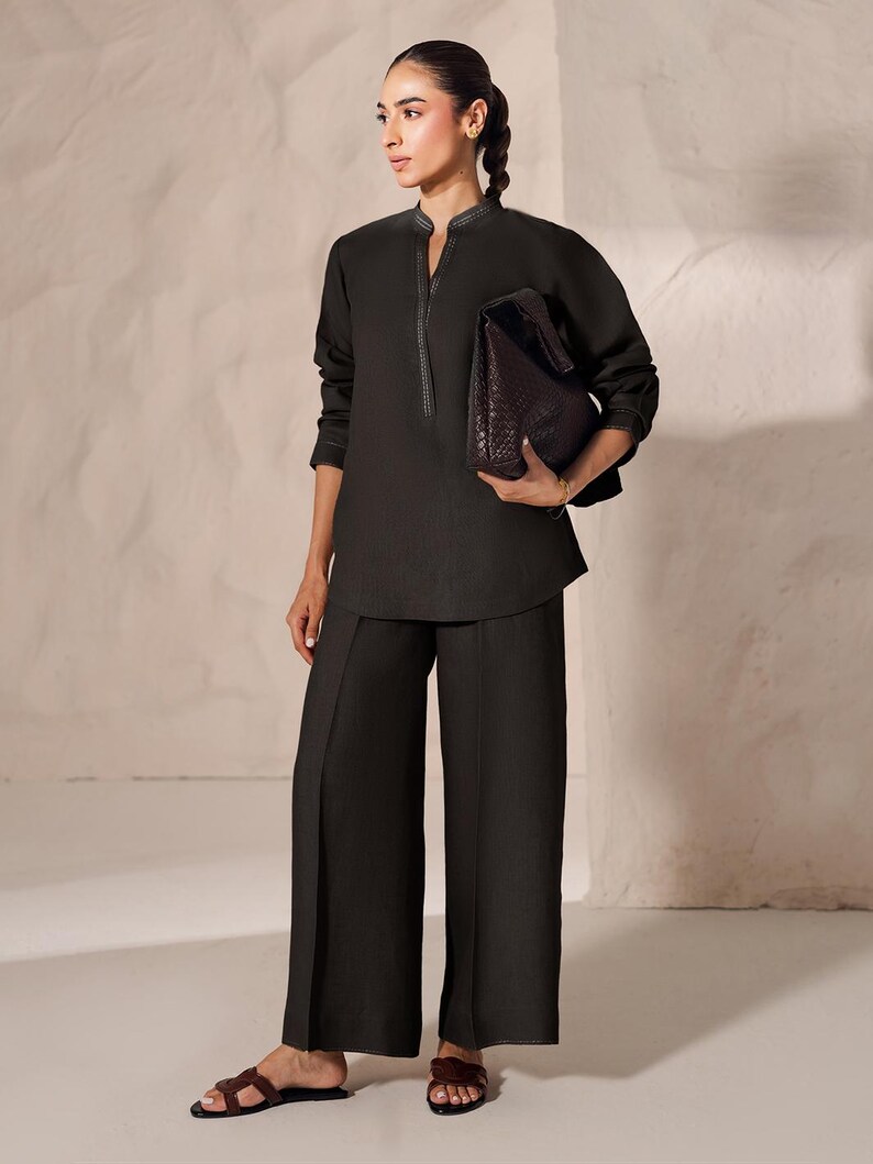 May include: A dark charcoal-colored ensemble featuring a long-sleeved tunic with a mandarin collar and wide-leg pants. The tunic has decorative stitching along the collar and cuffs. A dark brown leather tote bag is held in the hand. The outfit is paired with brown sandals.