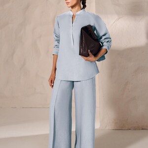 May include: A light blue linen outfit consisting of a long-sleeved tunic with a mandarin collar and wide-leg pants. A dark brown woven leather tote bag is held in the hand. The outfit is paired with brown leather sandals.