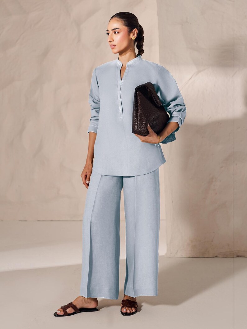 May include: A light blue linen outfit consisting of a long-sleeved tunic with a mandarin collar and wide-leg pants. A dark brown woven leather tote bag is held in the hand. The outfit is paired with brown leather sandals.
