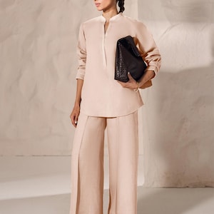 May include: A light peach-colored linen outfit consisting of a long-sleeved tunic and wide-leg pants. The outfit is paired with dark brown woven leather sandals and a dark brown woven leather tote bag.