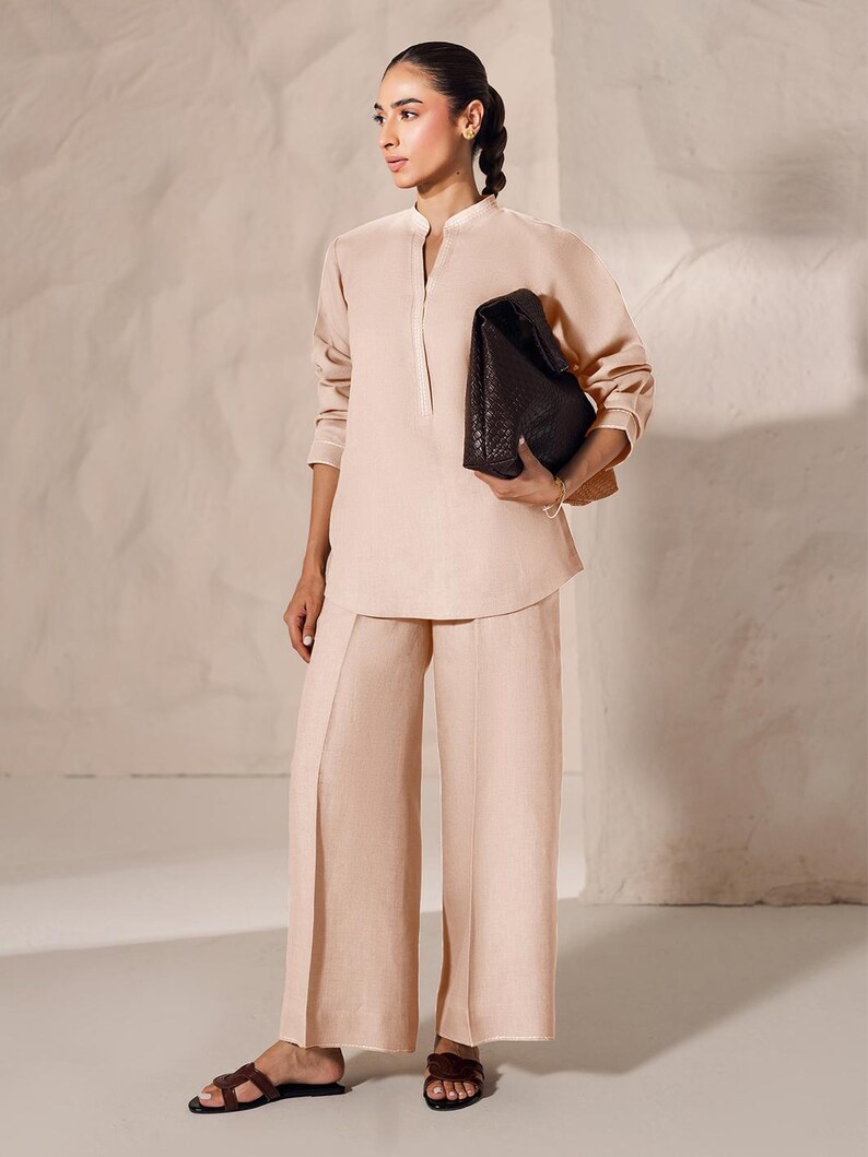 May include: A light peach-colored linen outfit consisting of a long-sleeved tunic and wide-leg pants. The outfit is paired with dark brown woven leather sandals and a dark brown woven leather tote bag.