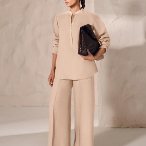May include: A woman wearing a beige linen outfit, including a long-sleeved tunic and wide-leg pants. She is holding a dark brown, woven leather handbag. The outfit is paired with brown sandals. The background is a neutral, textured wall.