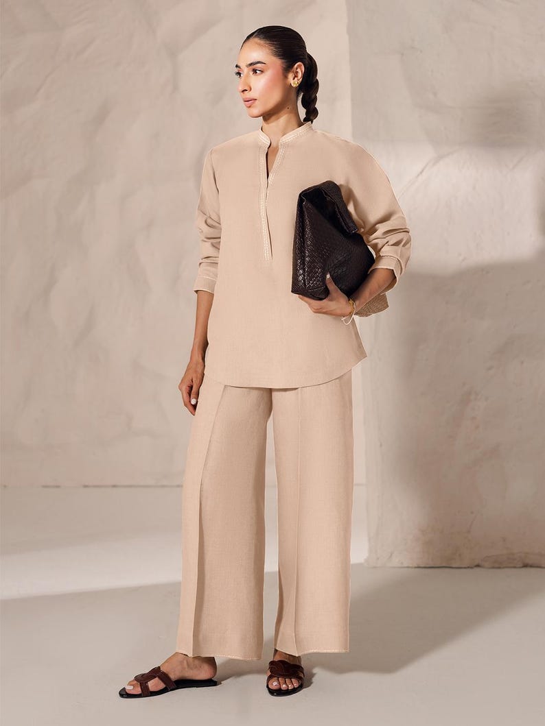 May include: A woman wearing a beige linen outfit, including a long-sleeved tunic and wide-leg pants. She is holding a dark brown, woven leather handbag. The outfit is paired with brown sandals. The background is a neutral, textured wall.