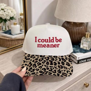 May include: White baseball cap with the embroidered phrase "I could be meaner" in red. The brim features a tan and black leopard print. A stylish accessory for casual wear.