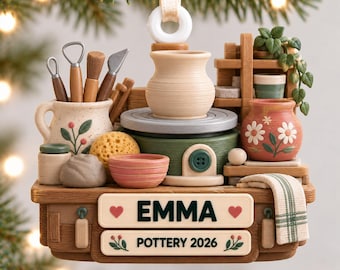 May include: A decorative ornament featuring a miniature pottery studio scene with a potter's wheel, clay tools, a small vase, and a potted plant. The base includes two rectangular plaques with the text EMMA and POTTERY 2026, accented by hearts and floral motifs.