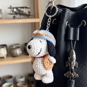 May include: A crocheted Snoopy keyring. The white and black Snoopy figure is dressed in a blue aviator cap and a tan vest. The keyring is attached to a black leather bag with a silver rocket charm.