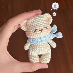 Pode incluir: Um pequeno urso de pelúcia de crochê em fio creme, usando um cachecol azul claro. O urso tem olhos fechados, detalhes em rosa nas orelhas e bochechas e uma boca pequena costurada. Duas bolhas de pensamento acima do urso mostram um coelho. O urso é segurado em uma mão.