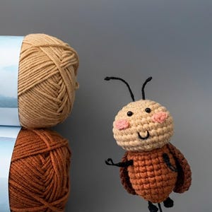 May include: A stack of two yarn skeins in shades of brown and tan, with a crocheted bee toy. The bee has a tan head with pink cheeks, a brown body, and black antennae and legs. The yarn is in a light blue label.