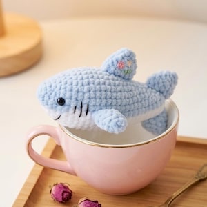 May include: A light blue crocheted shark toy with a white belly and black details, sitting in a pink teacup. The shark has a small flower detail on its fin. The teacup is on a wooden tray with dried rose buds and a gold spoon.