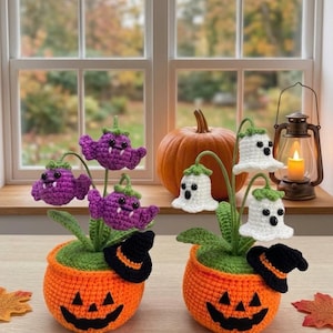 Pode incluir: Duas plantas de crochê em vasos laranja em forma de abóbora com rostos pretos. Um vaso apresenta flores roxas em forma de morcego com presas brancas, e o outro flores brancas em forma de fantasma com olhos pretos. Cada vaso tem um pequeno chapéu.