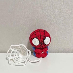 May include: A small, round, crocheted figure featuring a red head with black web-like stitching and large white eyes. The body is red with blue accents on the sides. A white, circular, crocheted spider web is attached to the figure by a white yarn string.