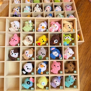 Puede incluir: Un organizador de madera lleno de coloridos llaveros de animales de ganchillo. Los llaveros incluyen una variedad de animales como una oveja, un pato, un oso y un conejo. Cada llavero se muestra individualmente en un pequeño compartimento, con los números 1-23 visibles.