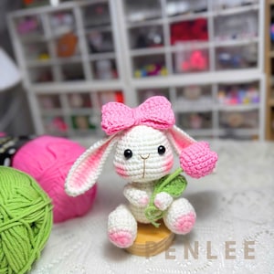 May include: A handmade crochet bunny with a pink bow, holding a pink flower and green leaves. The bunny is white with pink accents on the ears and feet. The bunny is sitting on a small wooden base.