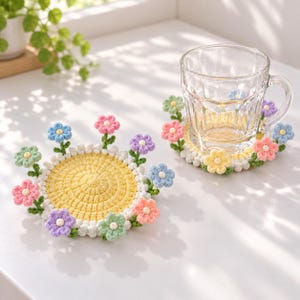 Può includere: Due sottobicchieri rotondi all'uncinetto con centro giallo, bordo bianco e piccoli fiori tridimensionali in tonalità pastello rosa, blu, verde e viola fissati sui bordi. Un sottobicchiere sostiene una tazza di vetro trasparente.