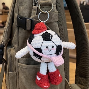 Peut inclure: Un porte-clés fait main en forme de ballon de football crocheté, avec un visage souriant, portant un chapeau rouge, une écharpe rose et des chaussures rouges. La balle noire et blanche a des bras et des jambes blancs. Accroché à un porte-clés argenté, il est suspendu à un sac à dos vert.