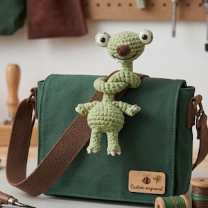 Pode incluir: Uma bolsa mensageiro de lona verde com uma alça marrom e uma figura alienígena verde de crochê anexada. A bolsa tem uma etiqueta de madeira que diz "Custom-engraved". O alienígena tem olhos grandes e um nariz marrom.