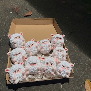 May include: A cardboard box filled with eleven white crochet sheep toys. Each sheep has pink accents on its ears and face. The toys are arranged on white paper, with a few leaves visible around the box.