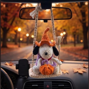 May include: A crochet hanging ornament featuring a white dog character wearing an orange witch hat and a purple cape, sitting on a small beige swing. The character holds a miniature broom and a small orange pumpkin, suspended by thick beige cords.