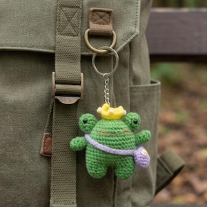 May include: A green crocheted frog keychain with a yellow crown and a purple sash, hanging from a silver chain. The frog has black eyes and a small purple pouch. The keychain is attached to a green canvas backpack.