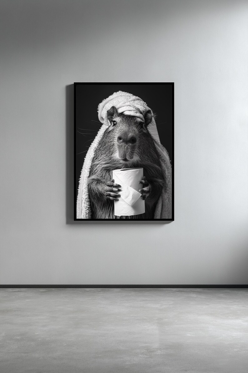 May include: A monochrome framed print featuring a capybara, wrapped in a towel and holding a roll of toilet paper. The capybara is the main subject, with the towel covering its head and shoulders. The background is dark.