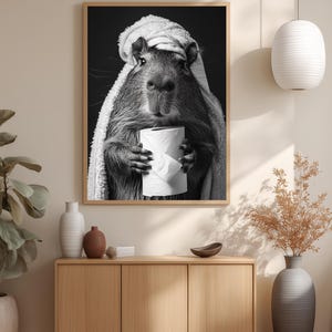 May include: Monochrome framed print featuring a capybara in a towel, clutching a toilet roll. The artwork hangs above a wooden cabinet with vases and a bowl. A white pendant light is suspended from the ceiling.