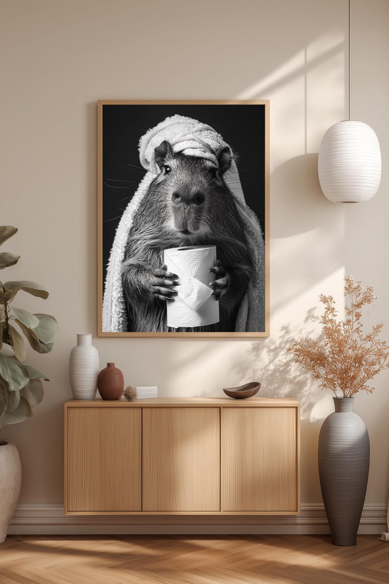 May include: Monochrome framed print featuring a capybara in a towel, clutching a toilet roll. The artwork hangs above a wooden cabinet with vases and a bowl. A white pendant light is suspended from the ceiling.
