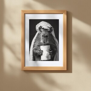 May include: Monochrome framed print of a capybara wrapped in a towel, clutching a roll of toilet paper. The image is set against a dark background, framed in light wood. A humorous and quirky piece of art.