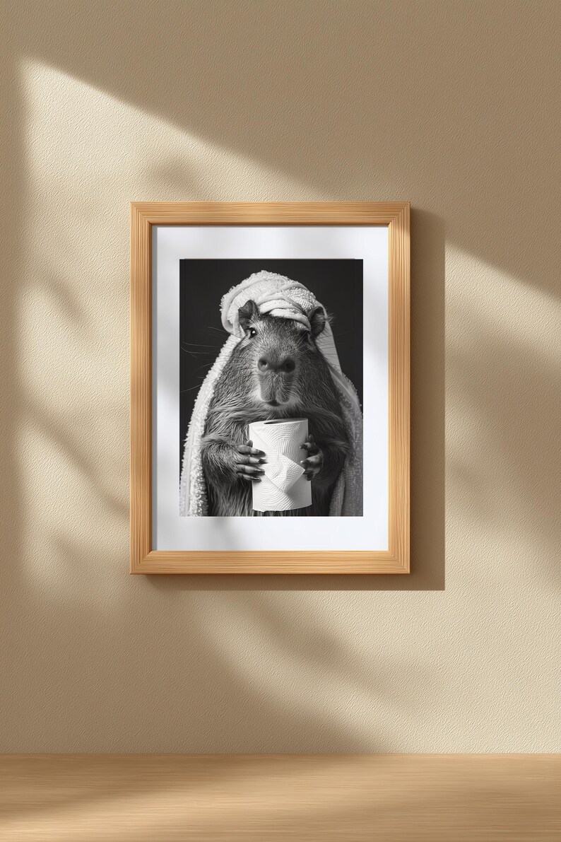 May include: Monochrome framed print of a capybara wrapped in a towel, clutching a roll of toilet paper. The image is set against a dark background, framed in light wood. A humorous and quirky piece of art.