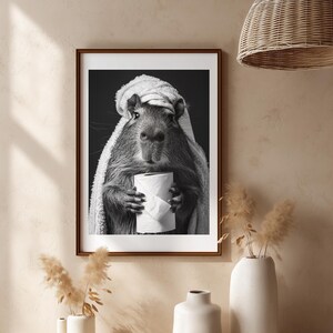 May include: Monochrome framed print of a capybara, wrapped in a towel and holding a roll of toilet paper. The image has a dark background. The print is displayed on a neutral wall, with decorative vases and a woven pendant light, creating a minimalist aesthetic.