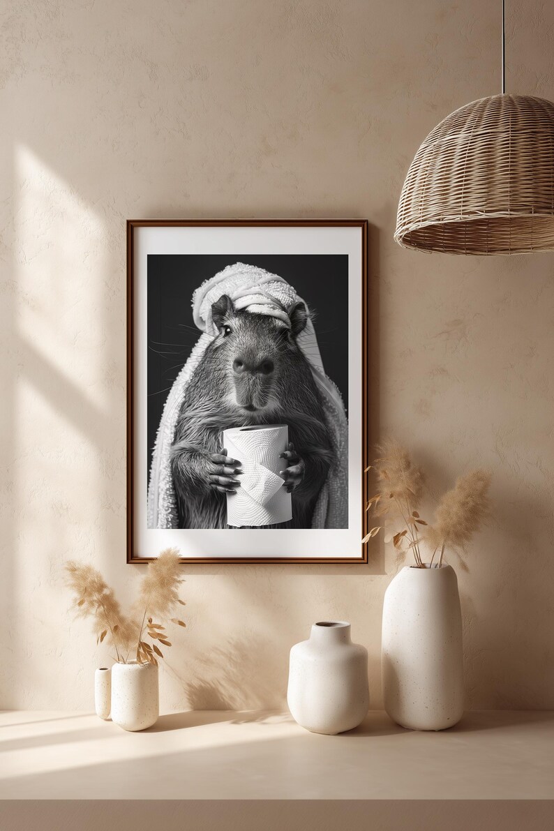 May include: Monochrome framed print of a capybara, wrapped in a towel and holding a roll of toilet paper. The image has a dark background. The print is displayed on a neutral wall, with decorative vases and a woven pendant light, creating a minimalist aesthetic.
